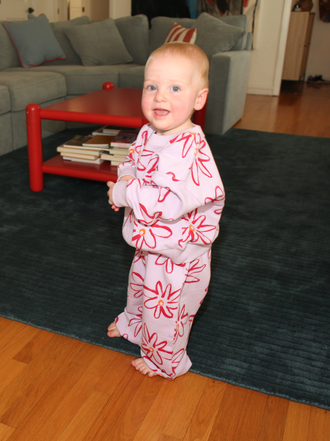 A toddler wearing the CHARLIE SWEAT SET in pink with a flower print stands barefoot on a hardwood floor beside a blue rug in the living room.