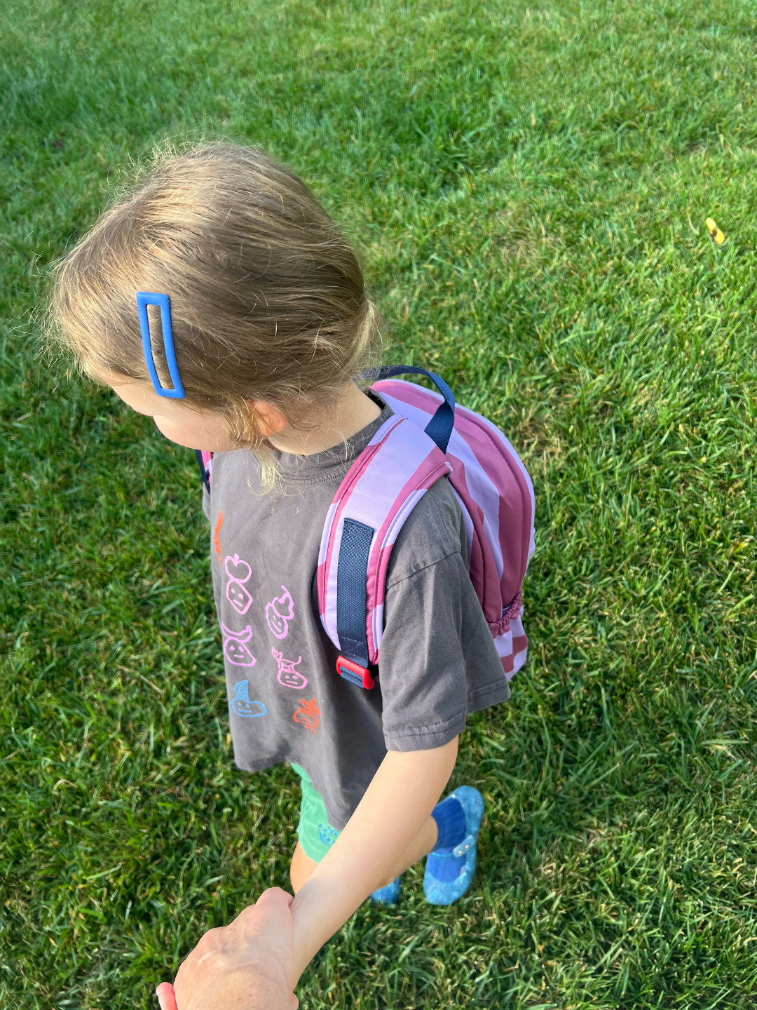 Purple Stripe | A young child walking outdoors on green grass, wearing a gray graphic t-shirt, blue sneakers, and a purple striped backpack.
