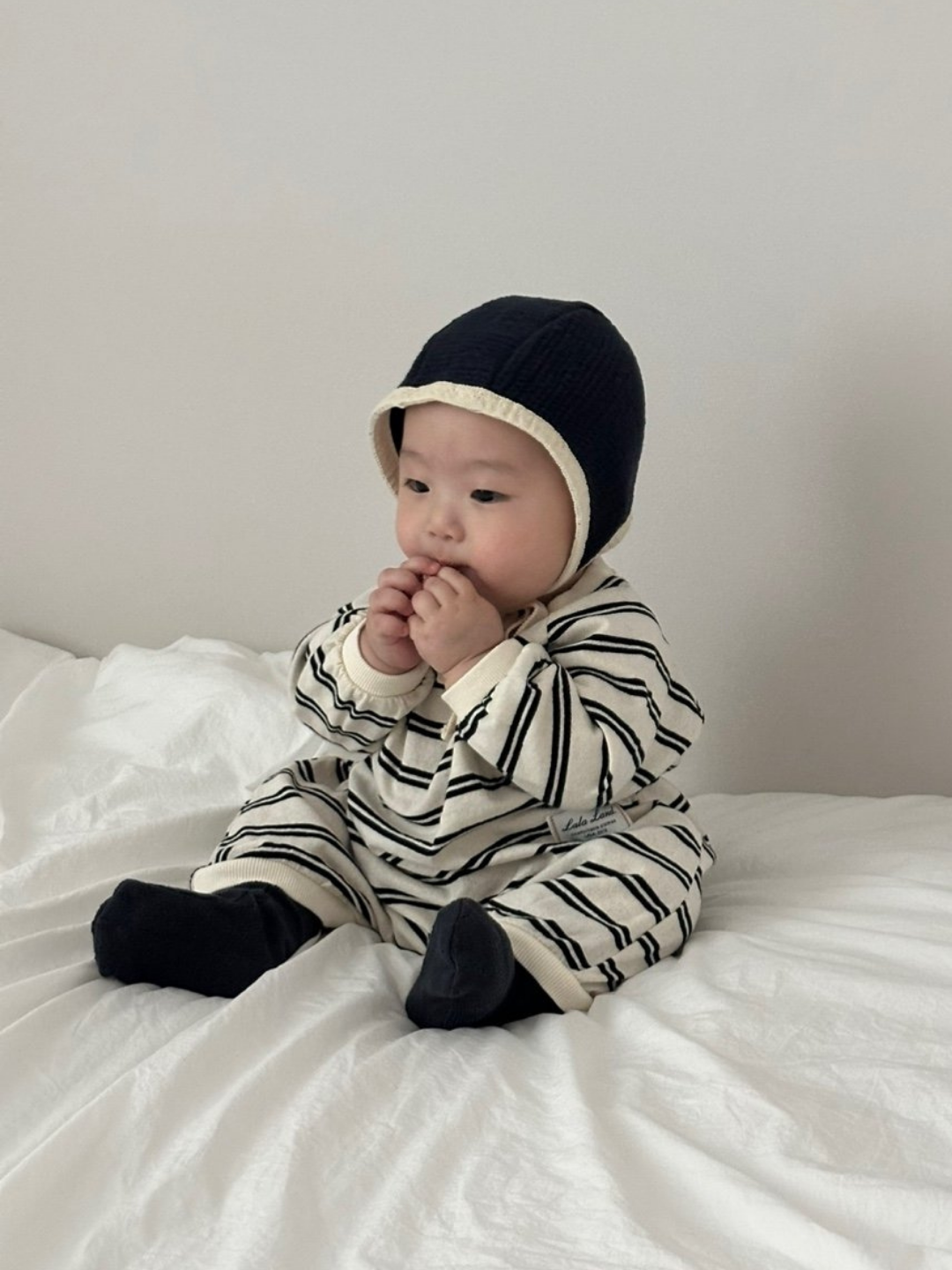 A baby wearing the SLOUCHY STRIPE ROMPER from a Korean brand and a dark bonnet sits on a white bed, hands near their mouth, gazing slightly to the side.