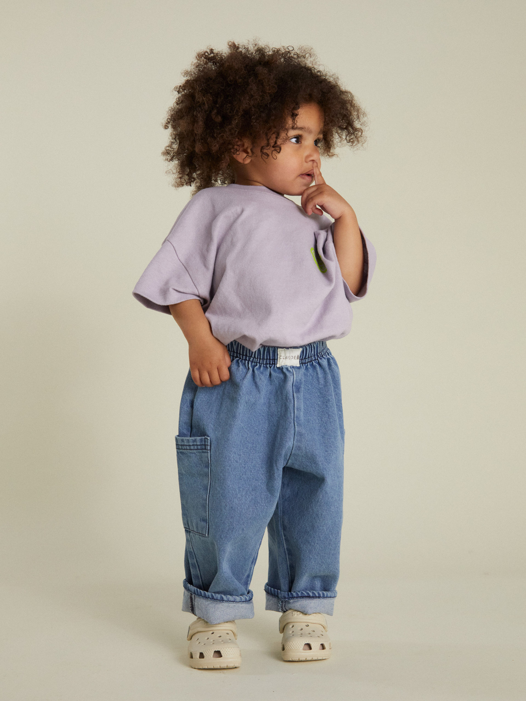 A young child with curly hair wears a loose lavender shirt, EMERSON DENIM BABY TROUSER with a side pocket, and light slip-on shoes, standing against a plain background.