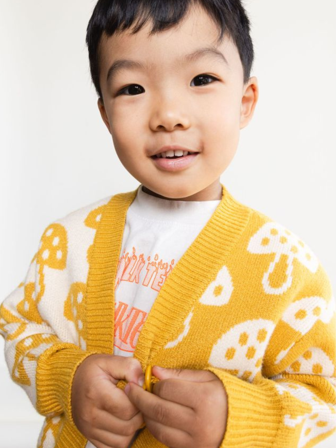 A young child smiles at the camera, wearing the SHROOM CARDIGAN—a white and yellow cardigan with a charming mushroom pattern—paired with a white T-shirt featuring an orange graphic. The description perfectly captures their joyful spirit.