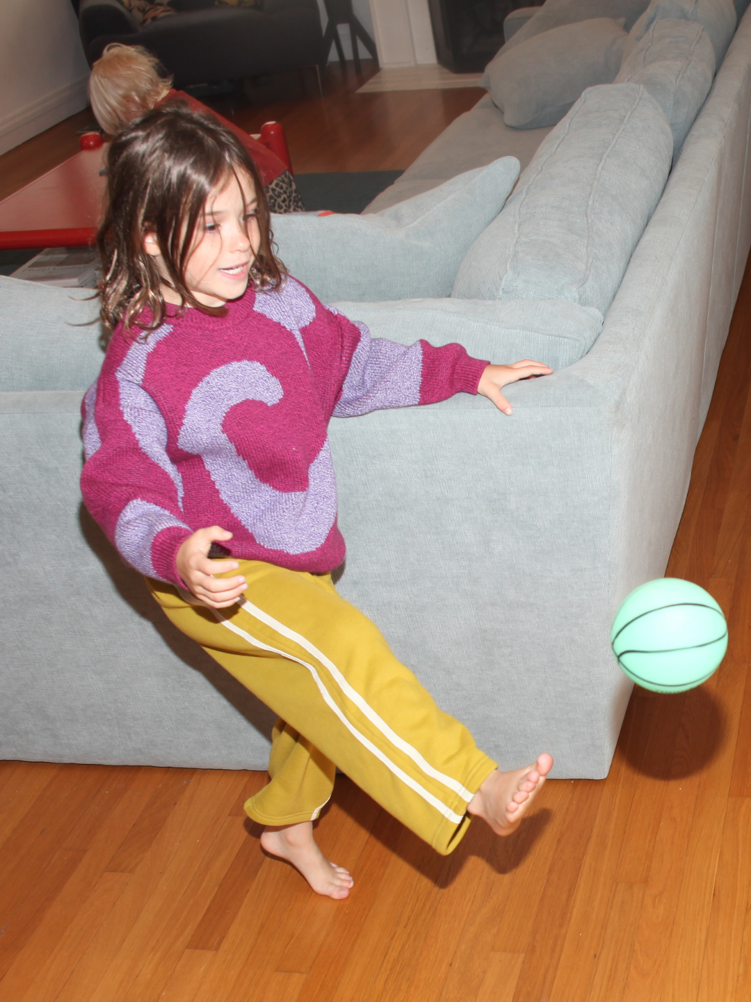 A child wearing SUNDAY TRACKPANTS in yellow and a purple-maroon sweater kicks a light green ball inside on a wooden floor, beside a light blue sofa.