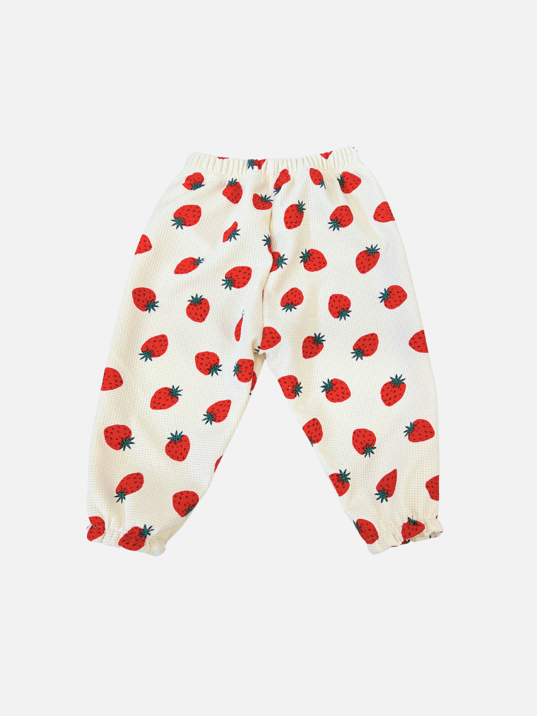 The STRAWBERRY WAFFLE JOGGER from a popular Korean kids brand features a charming red strawberry print on white waffle-textured fabric, complete with an elastic waistband and cuffs for comfort and style.