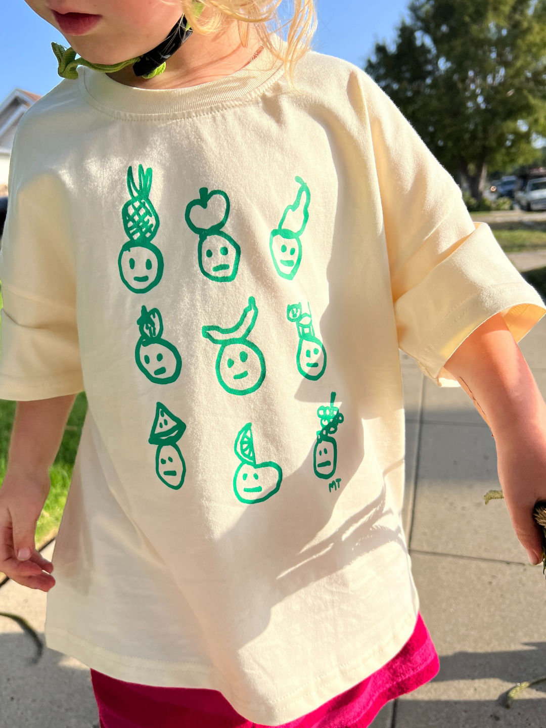 A child is wearing a cream Fruit Face tee with a bike helmet and pink shorts. 