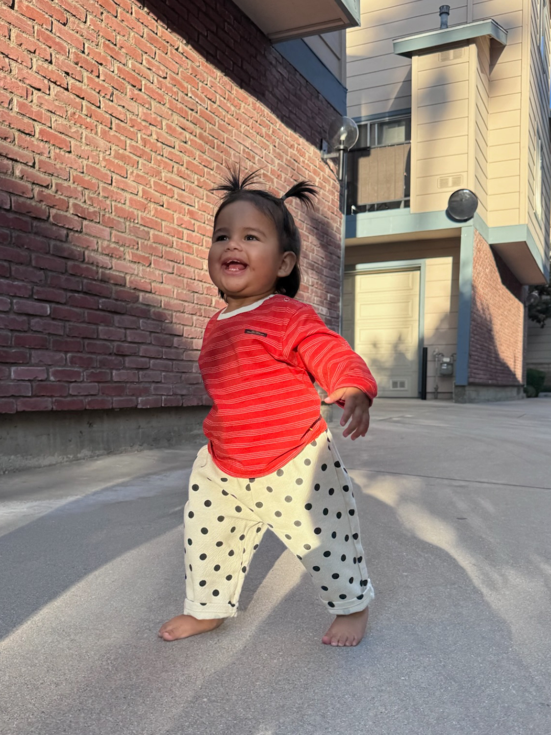 Child in red shirt and polka dot pants standing on a street with buildings in the background