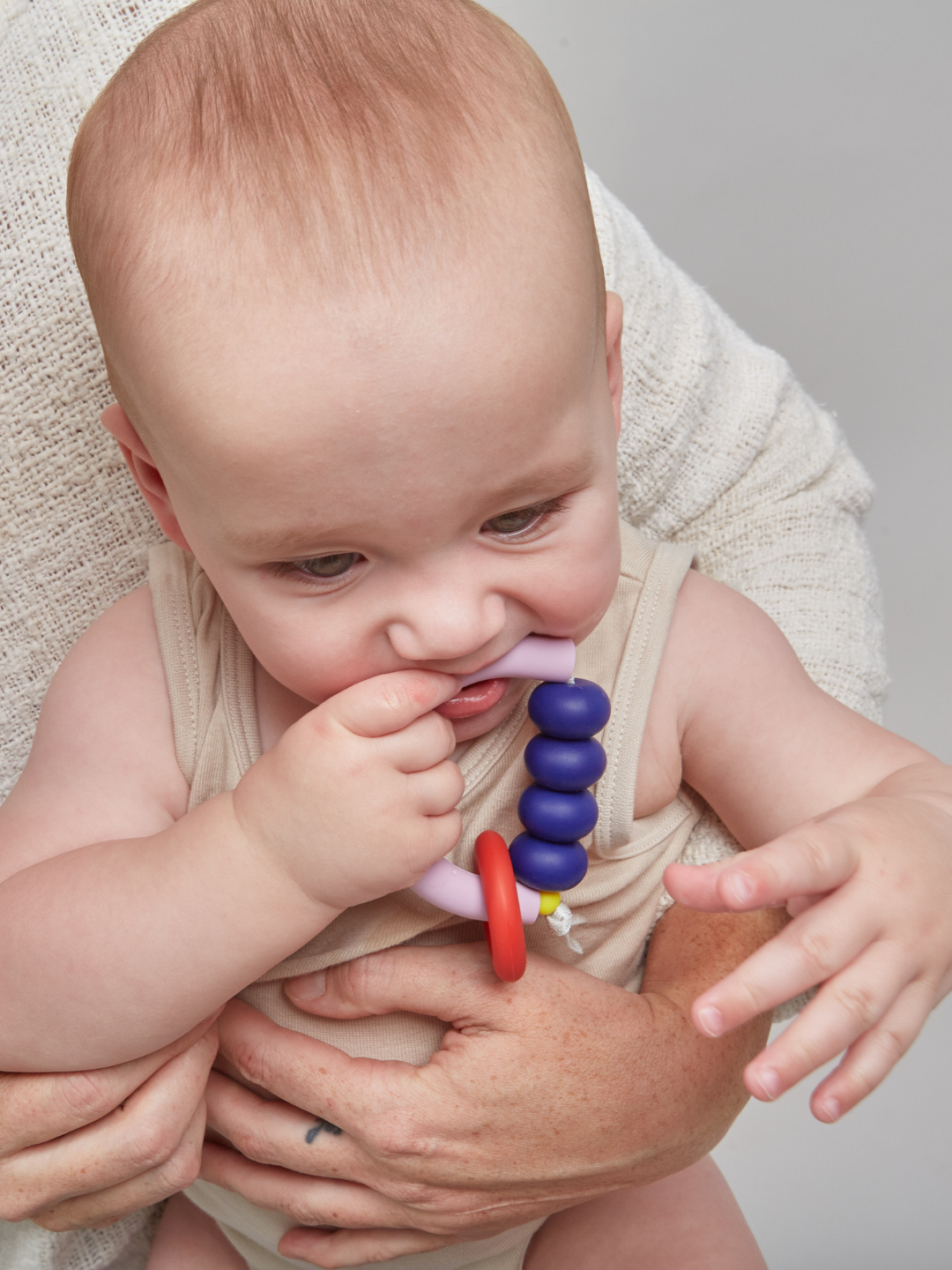 A baby in a beige outfit is held by an adult and chews on the MT X JM ARCH RING TEETHER, which features colorful purple and red beads and is a sensory-friendly option made in the USA.