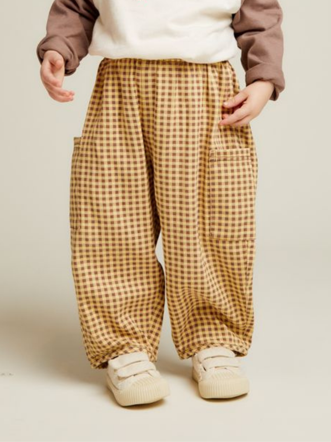 A child wearing EMERSON OCHRE GINGHAM TROUSER, paired with a cream and brown sweatshirt and cream shoes, stands against a plain background.