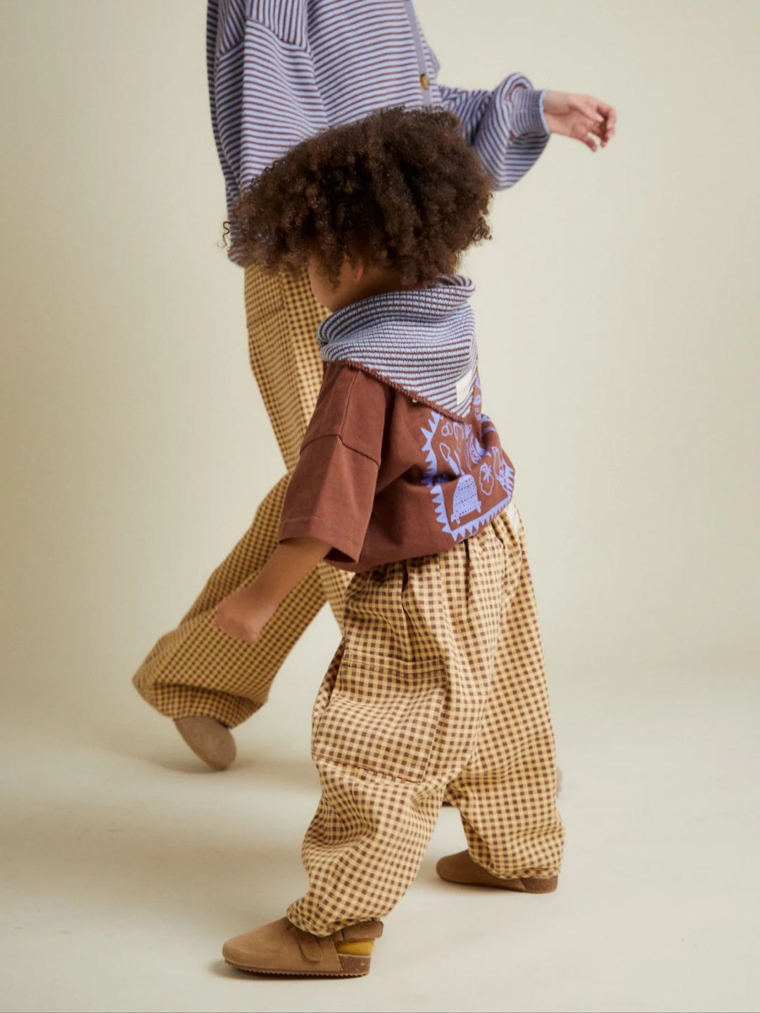 An adult and a child walk side by side, both wearing EMERSON OCHRE GINGHAM TROUSER pants. The child pairs them with a brown graphic-back shirt and a striped scarf.