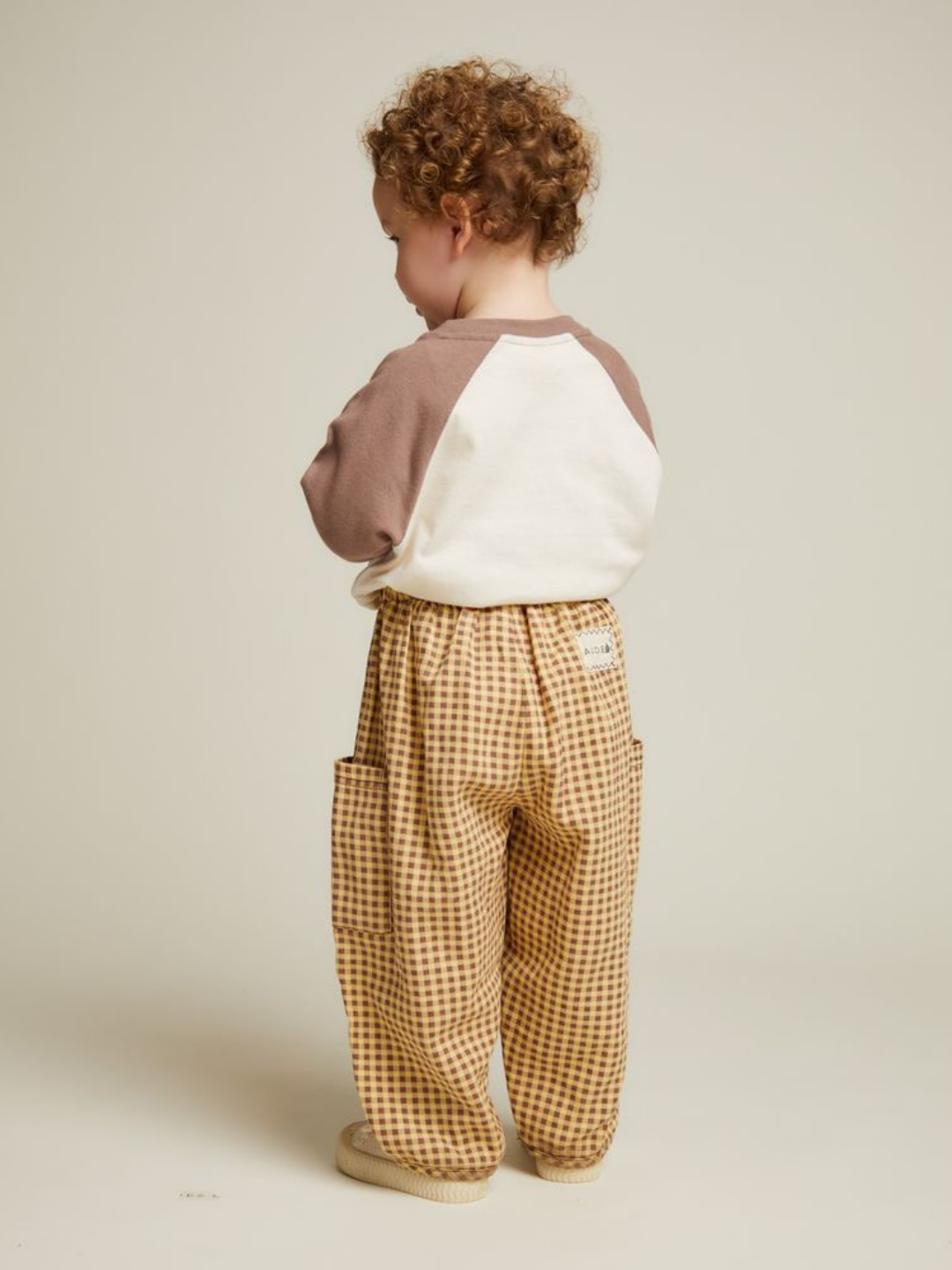 A young child with curly hair stands facing away, wearing a cream and brown long-sleeve shirt and EMERSON OCHRE GINGHAM TROUSER with pockets against a plain background.
