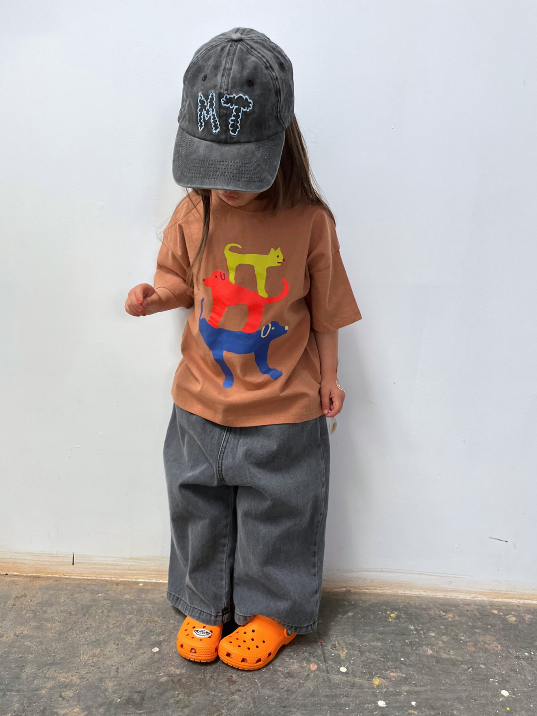Child wearing a brown t-shirt with colorful cat designs, gray cap, gray pants, and orange Crocs standing against a white wall.