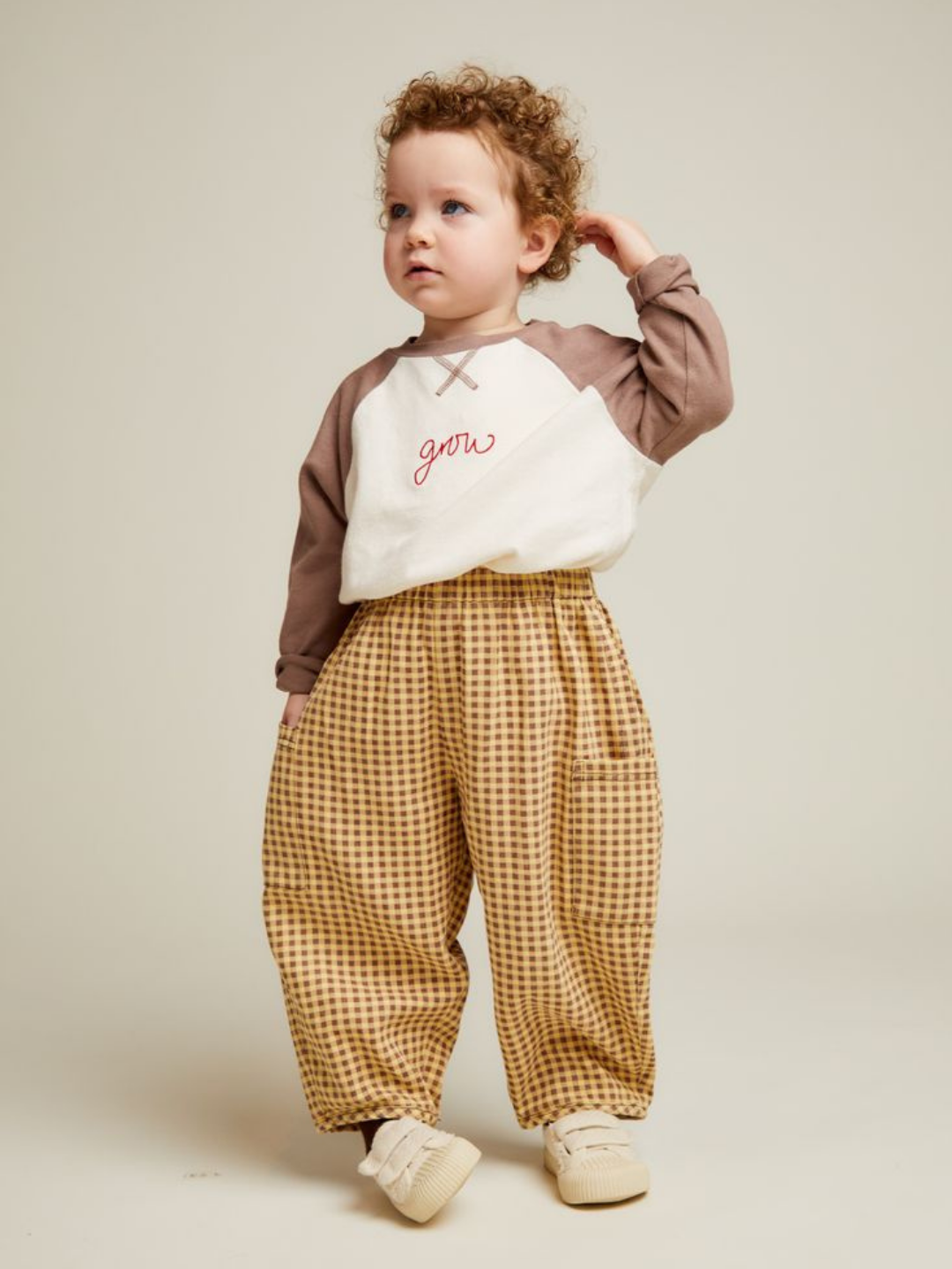 A young child with curly hair wears a cream and brown long-sleeve "grow" shirt, EMERSON OCHRE GINGHAM TROUSER, and beige shoes, standing against a plain background.