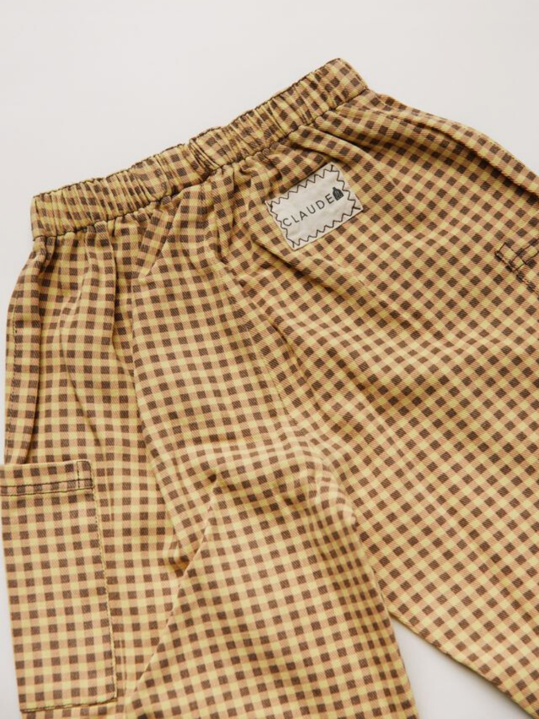 The EMERSON OCHRE GINGHAM TROUSER features brown and yellow checkered fabric, an elastic waistband, and a "CLAUDE" patch sewn near the back pocket.