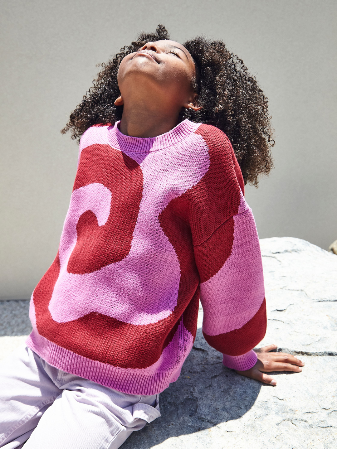 A girl wearing a kids crewneck sweater in bright pink, with a large single red swirl design that covers the entire sweater. She is seated on a rock, with her face turned upwards towards the sun. She wears light purple pants.