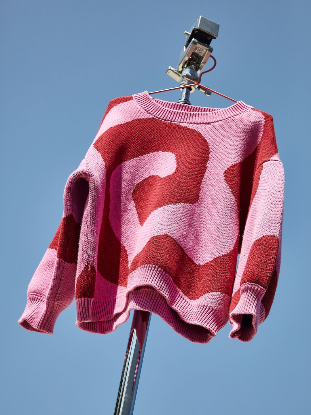 Front view of kids crewneck sweater in bright pink, with a large single red swirl design that covers the entire sweater. The sweater is shown on a hanger, hanging from a metal photography stand, against a blue sky.