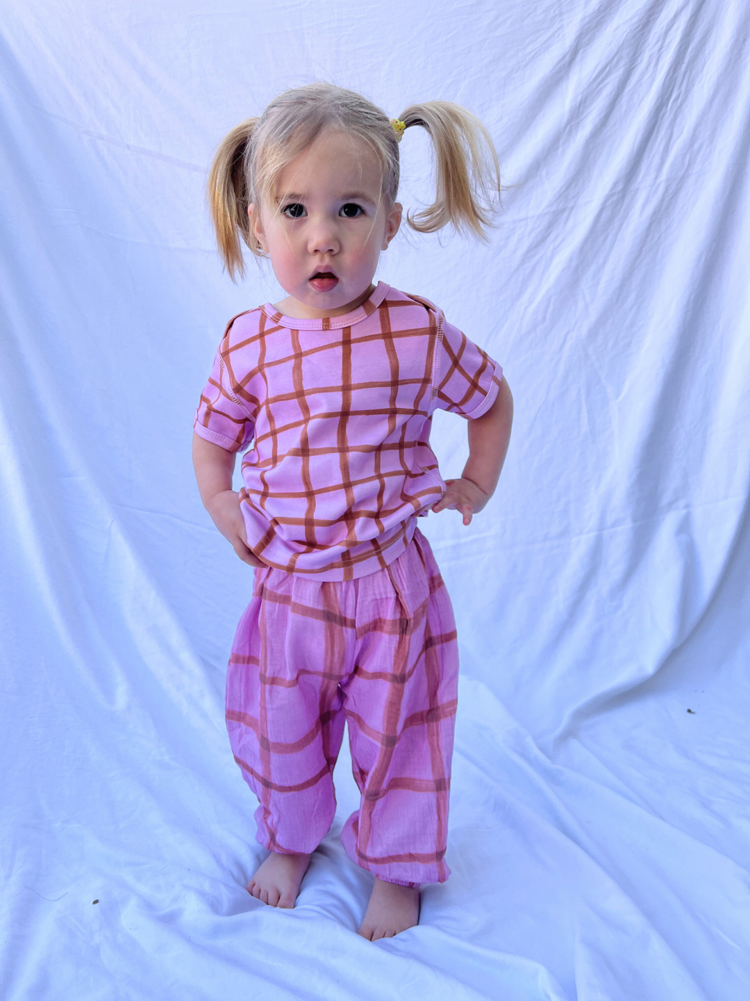 A barefoot toddler with pigtails stands against a white background, wearing the paintbrush check muslin pants in pink featuring a hand-drawn grid pattern.