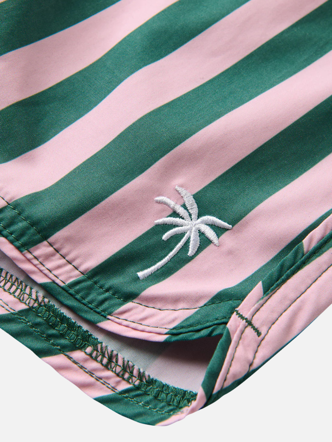 Close-up of RETRO FLARE BOARDSHORTS CABANA STRIPE, crafted from recycled polyester fabric with green and pink vertical stripes and a small white embroidered palm tree near the stitched hem.