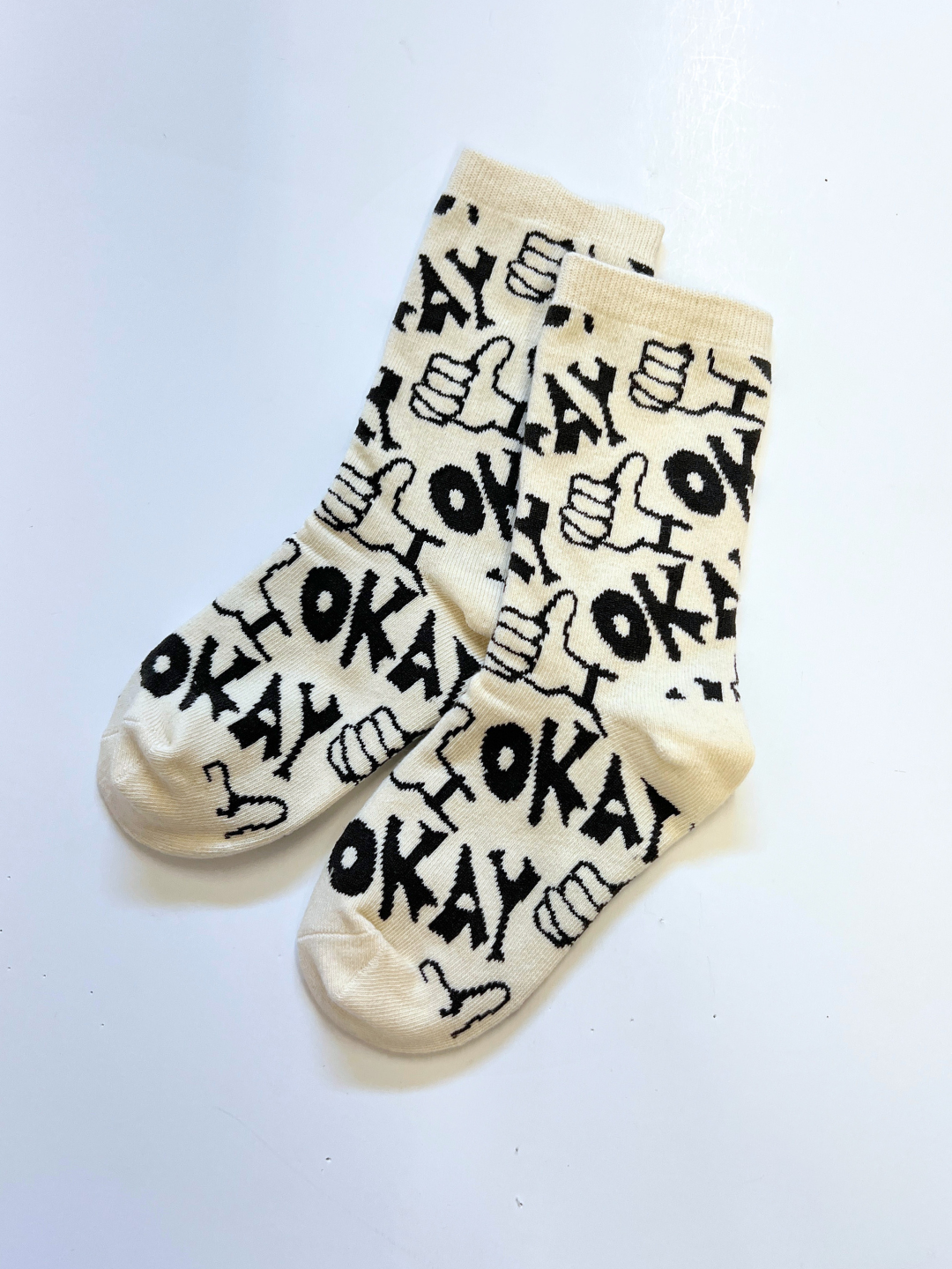 Ivory | The OKAY SOCK features a comfy, cream-colored design with black "OKAY" text and playful thumbs-up hand symbols in a hand-drawn pattern, displayed on a white surface.