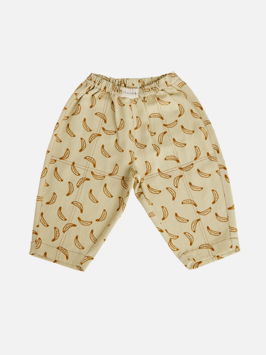 Image of The RIAN BANANA TROUSER is a beige unisex pant made from organic cotton twill with an elastic waistband and playful repeating banana print, featuring oversized balloon legs and shown laid flat on a white background.