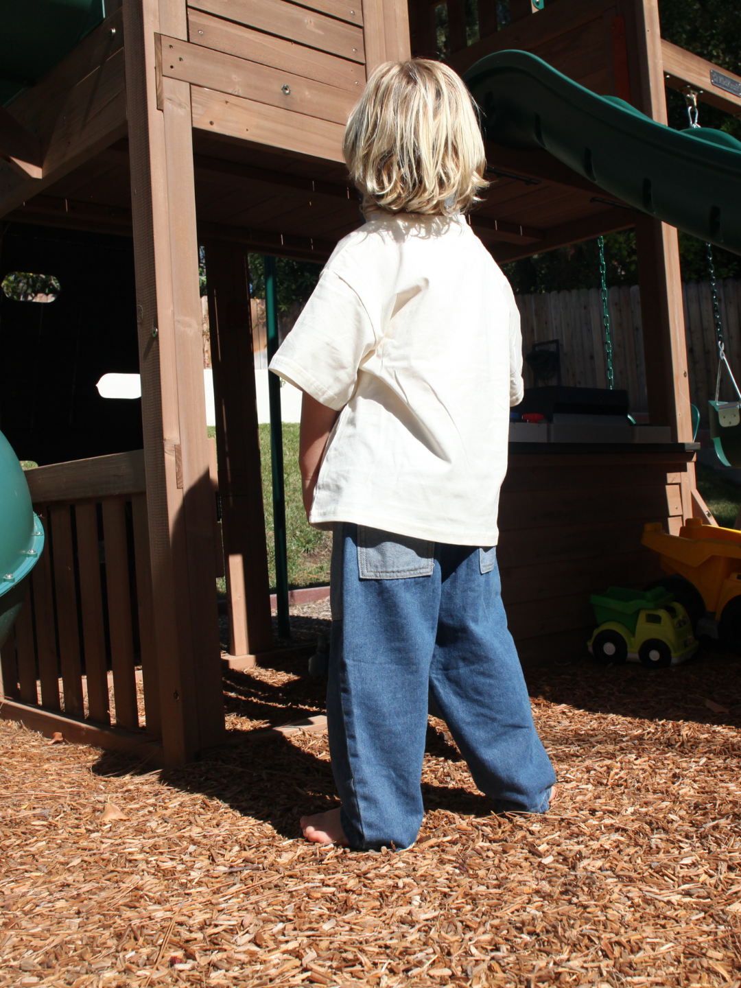 Child standing on a playground with wooden equipment and mulch below, wearing baggy blue jeans.