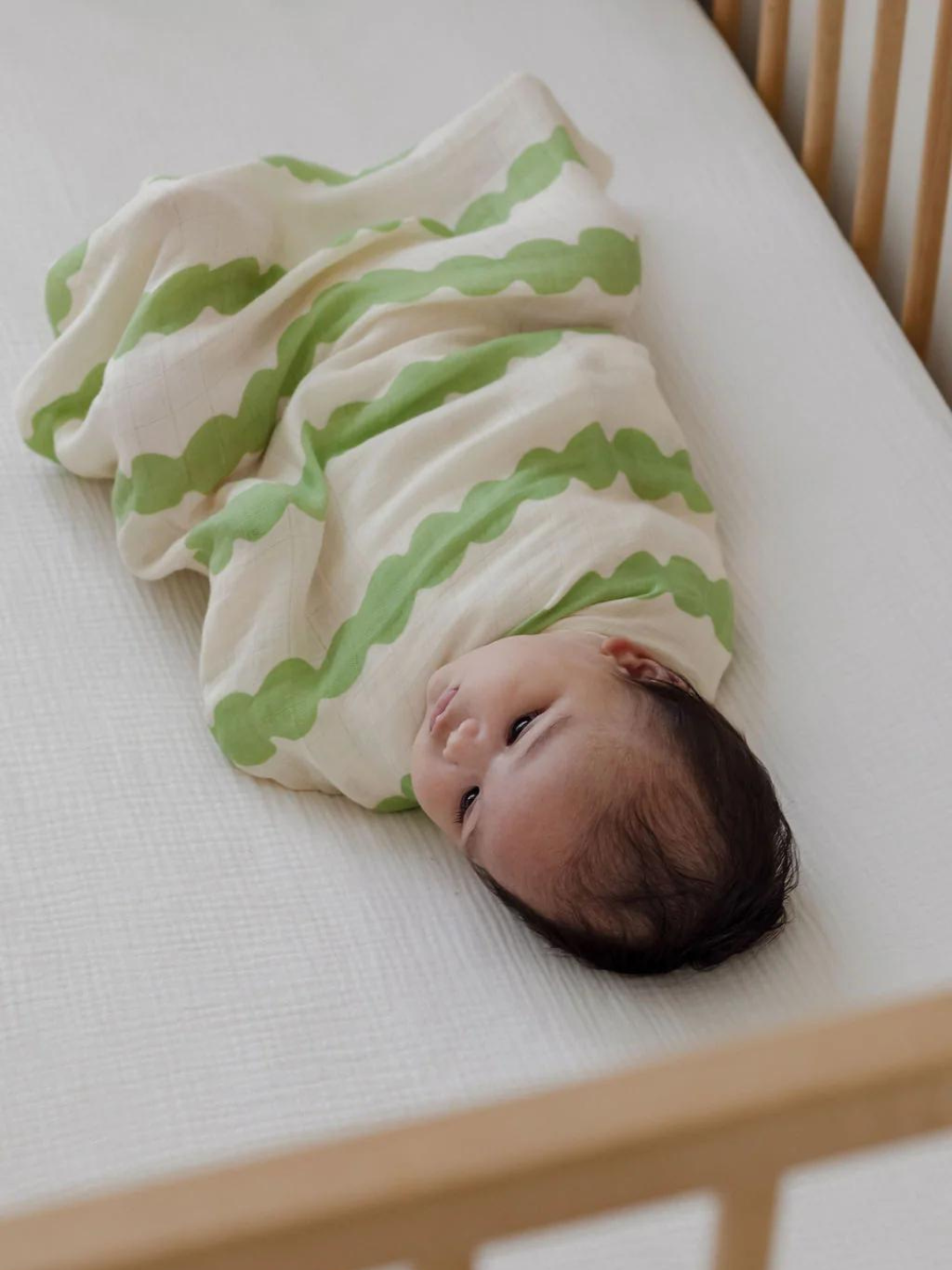 A baby wrapped in an ORGANIC BAMBOO SWADDLE WRAP lies on a white mattress inside a wooden crib.