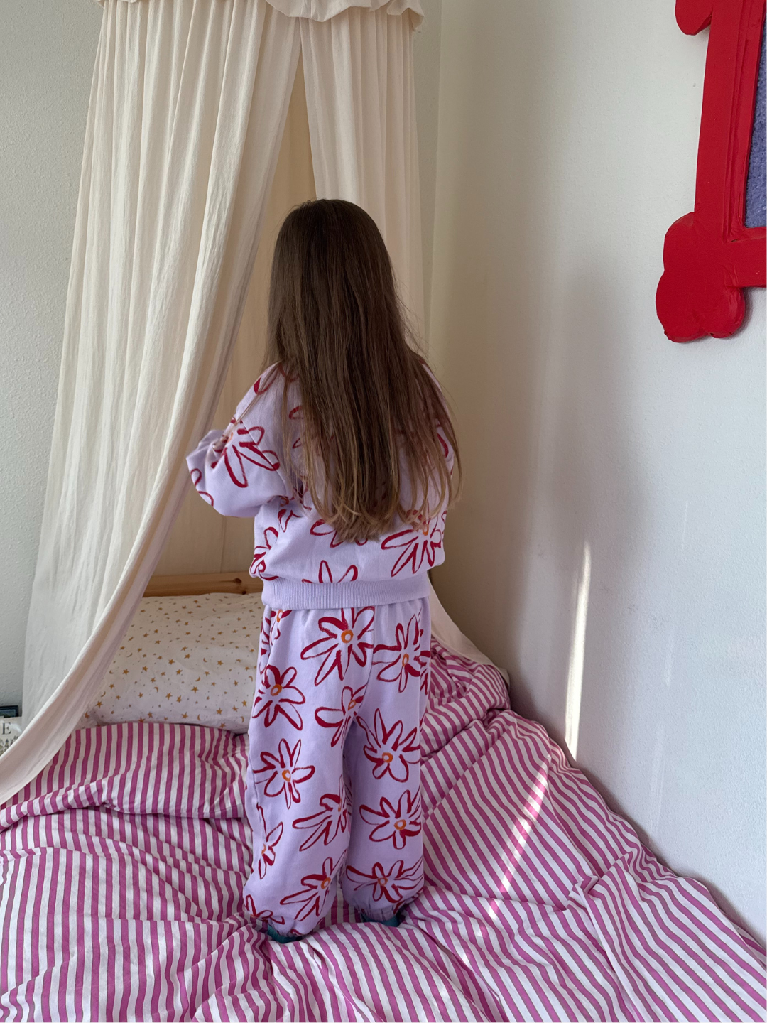 Lilac | Child in a floral outfit standing on a bed with striped bedding, looking out a window.