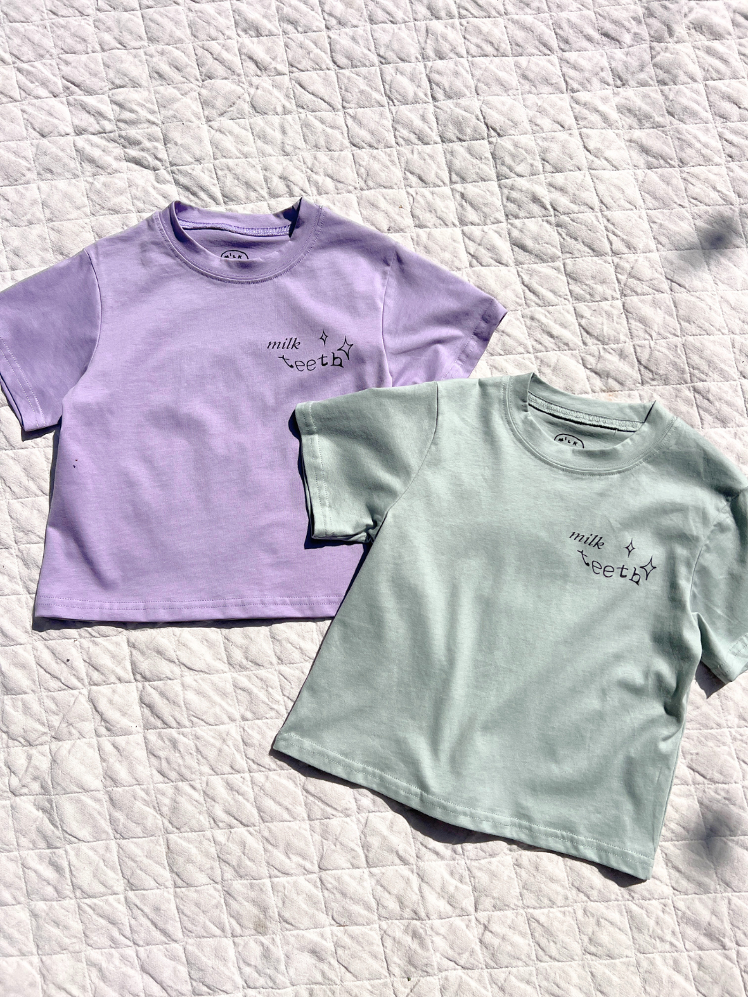 Two MILK TEETH TEE shirts in lavender and mint green, featuring "milk teeth" and star designs by artists, are laid flat on a quilted white surface.