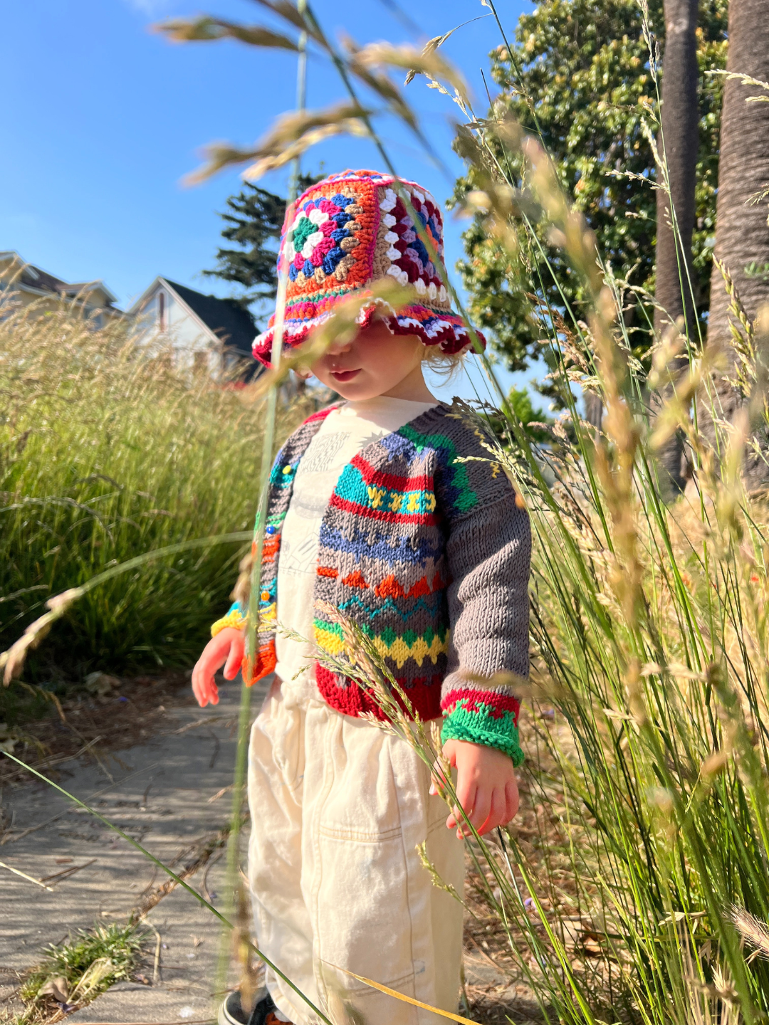 A young child wears the HAND-KNITTED COTTON CARDIGAN and stands in tall grass with green bushes and palm trees in the background on a sunny day.