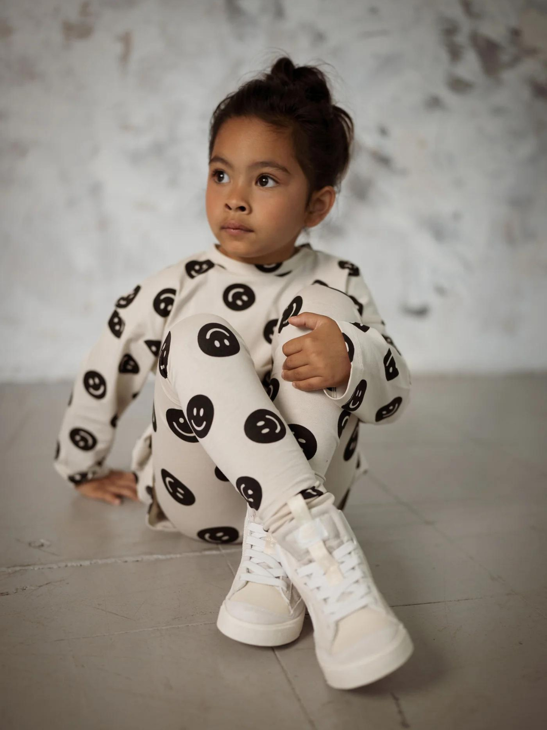 A front view of the cream colored leggings with a black smiley face pattern on a child sitting down.