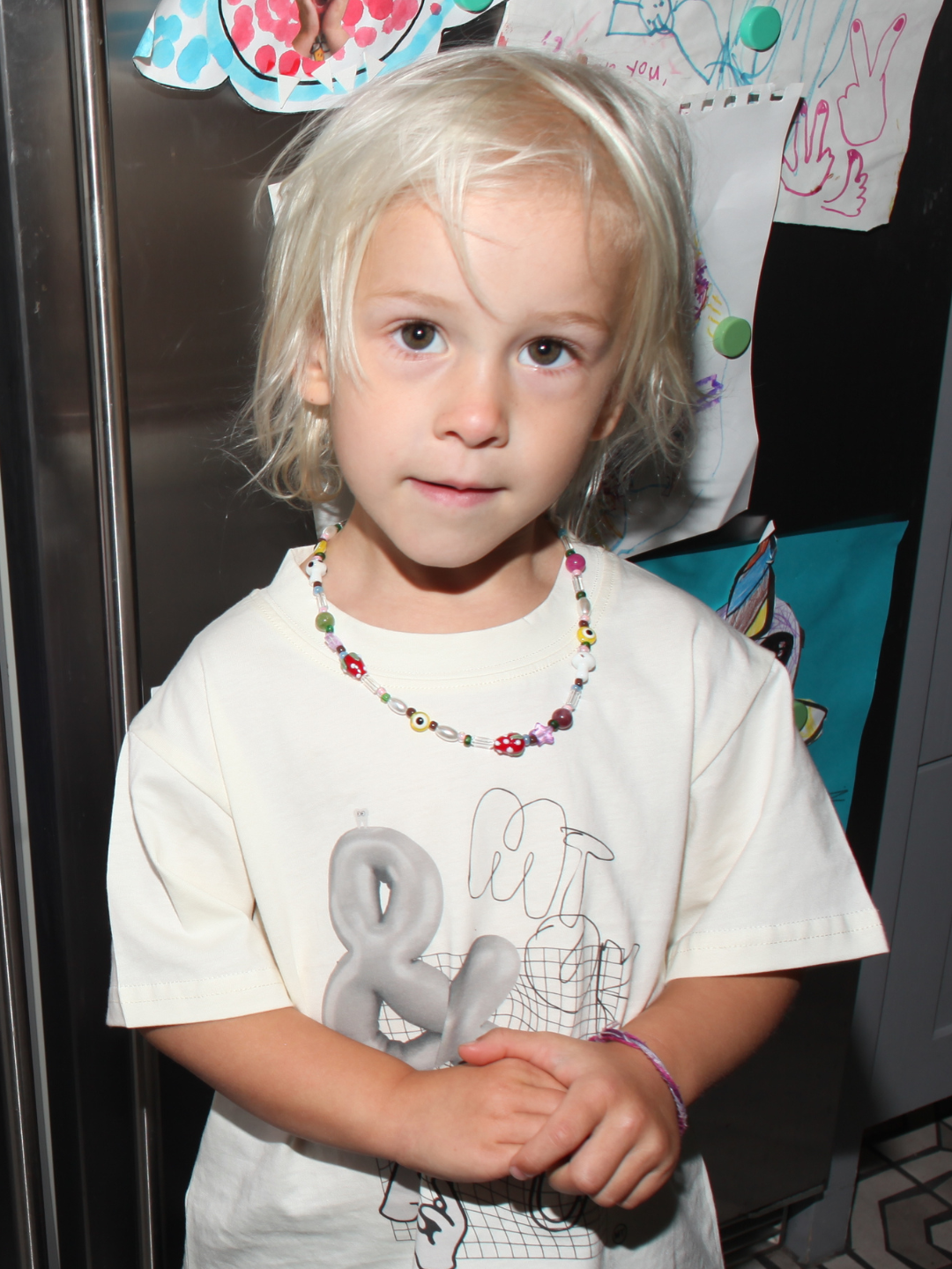 A young child with blond hair wearing the GAIA NECKLACE, which features handmade strawberry and mushroom beads, stands in front of a refrigerator covered in colorful children's drawings.