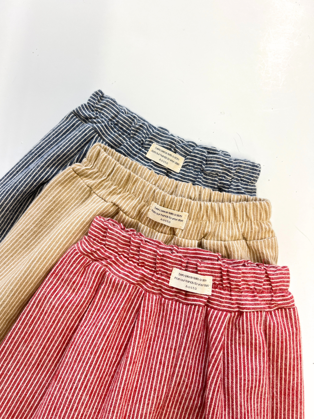 Three SOFT STRIPE BABY PANT pairs in blue, beige, and red with elastic waists are overlapped on a white surface, each showing the label on the waistband.