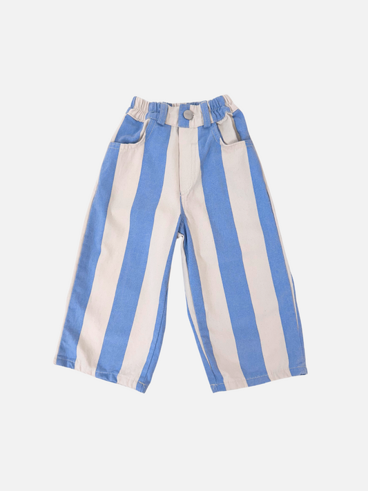 Image of The CABANA STRIPE PANTS are blue and white vertically striped children's pants from a Korean brand. They feature an elastic waistband, high waist, front button, and pockets. Made from durable cotton canvas, they lay flat on a plain white background.