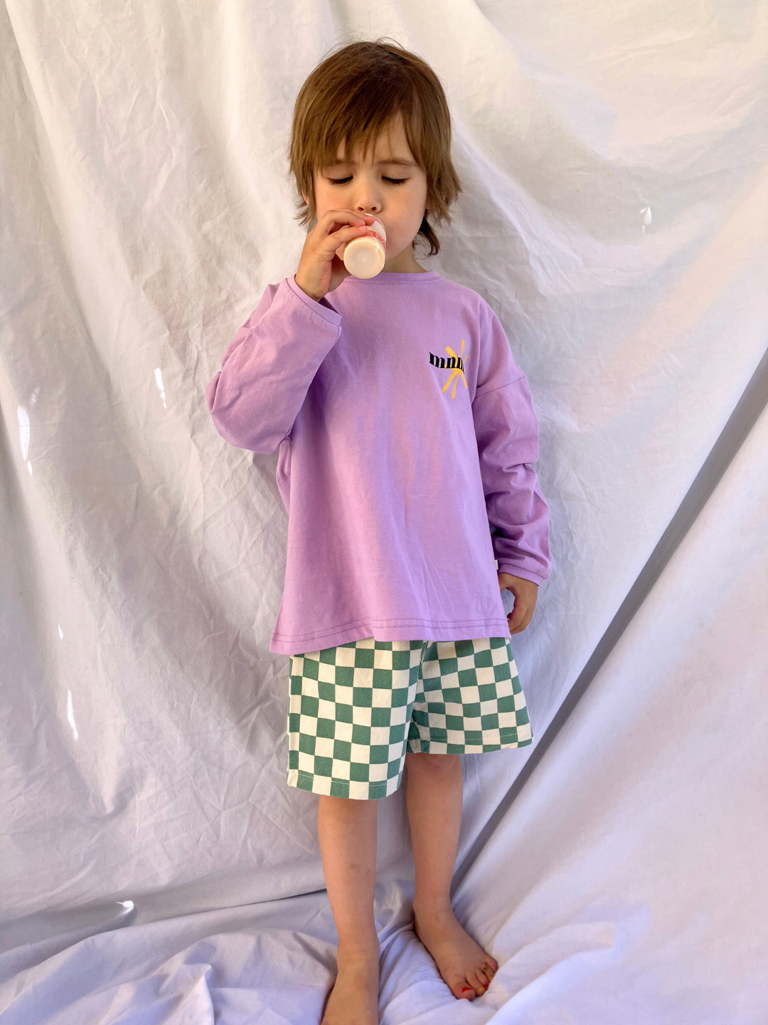 Purple | A child wearing the kids Minimal Longsleeve Tee in purple, paired with checkerboard patterned shorts in teal and ivory. 