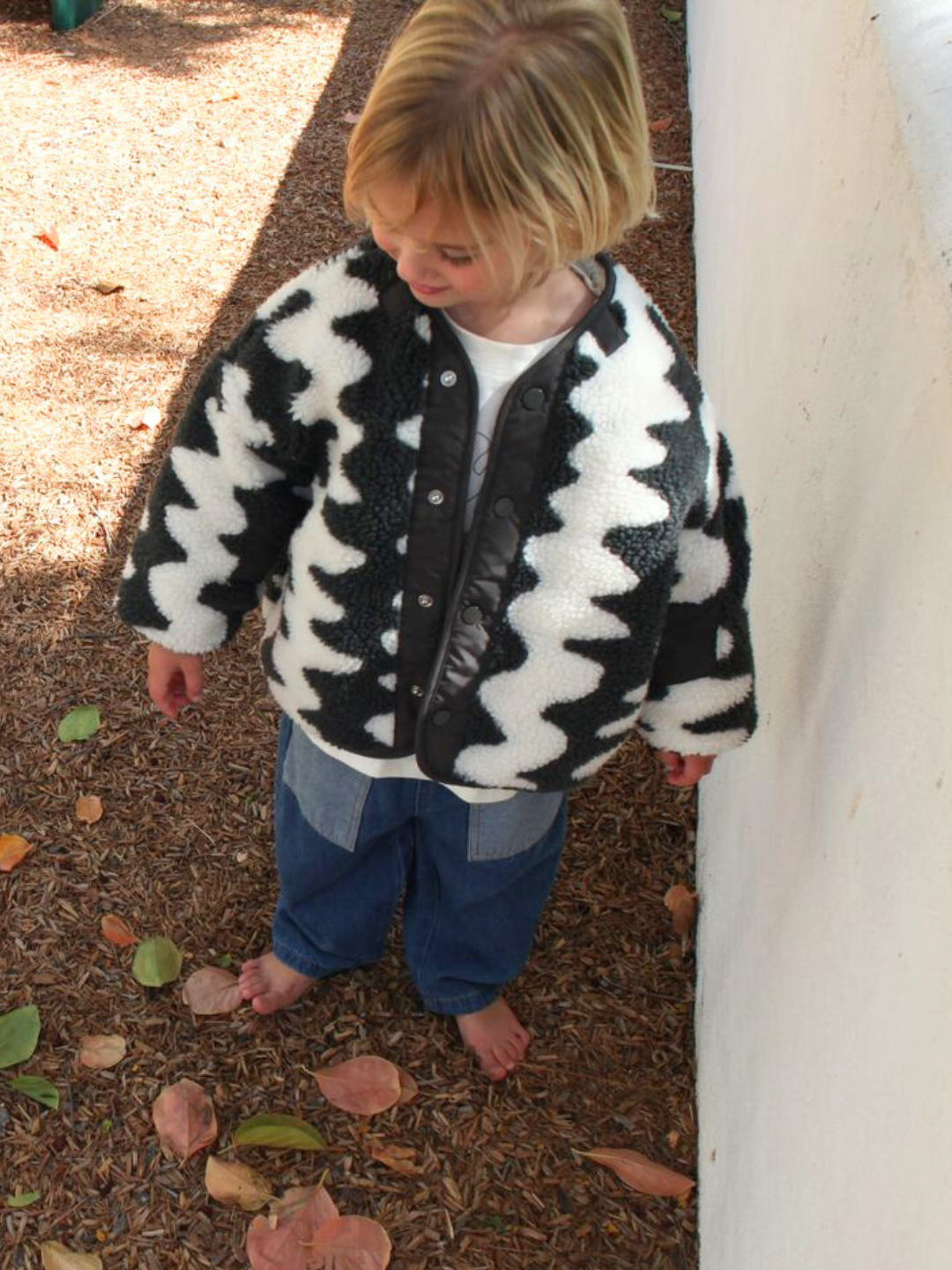A young blonde child stands barefoot on mulch, wearing loose blue jeans and the WAVELENGTH SHERPA JACKET, which features a black-and-white zigzag pattern and a cozy fleece lining.