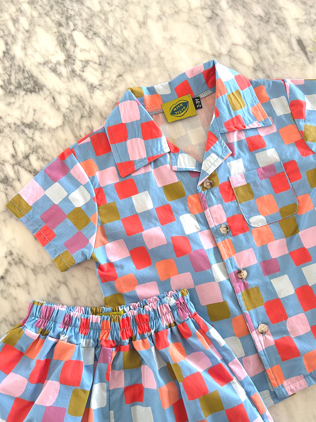 Blue | A kids' shirt and shorts set laid on a table, in a pattern of red, orange, pink, lilac and green squares on a blue background