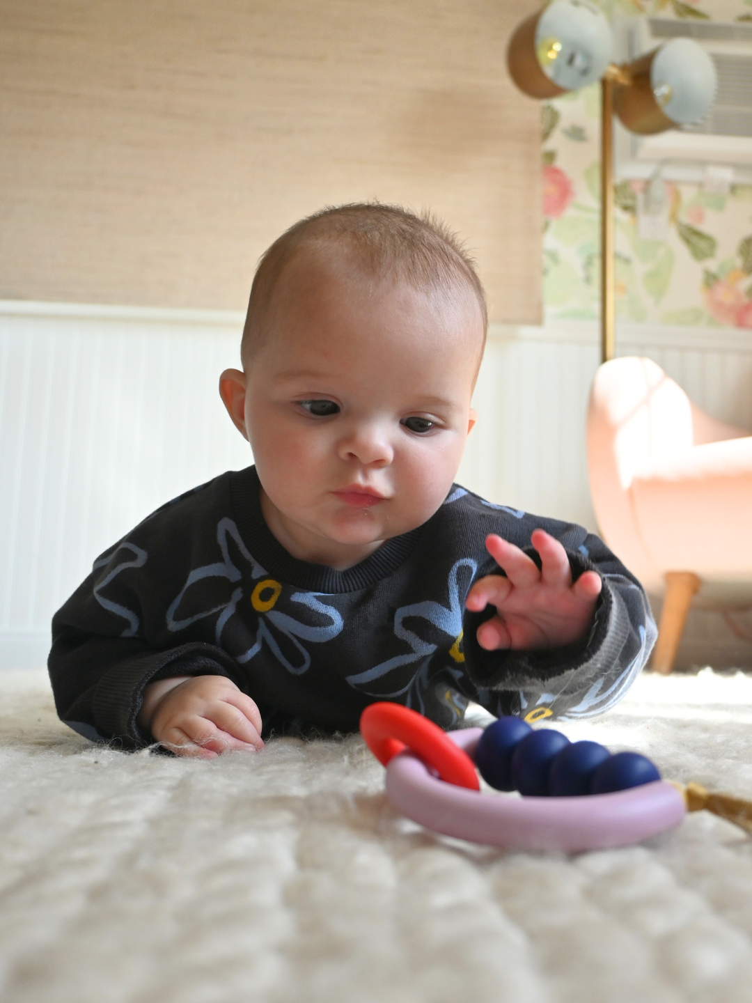 A baby lies on a soft, light rug, reaching for the MT X JM ARCH RING TEETHER in purple and red, with a chair and lamp visible in the background.