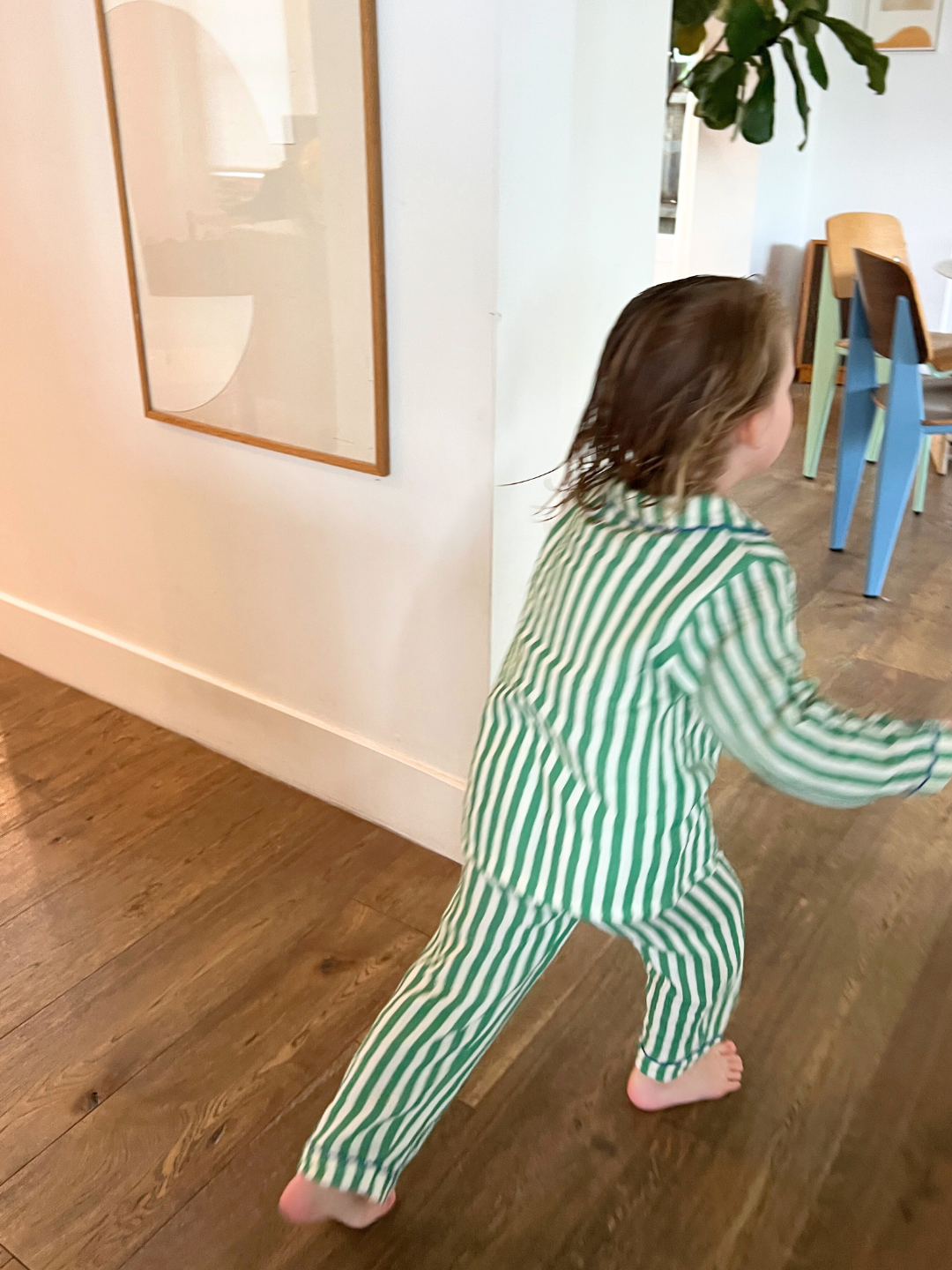 A child in the MOVIE NIGHT SET, featuring green and white striped set made from breathable cotton muslin, joyfully runs through a room with wooden floors and modern furniture.