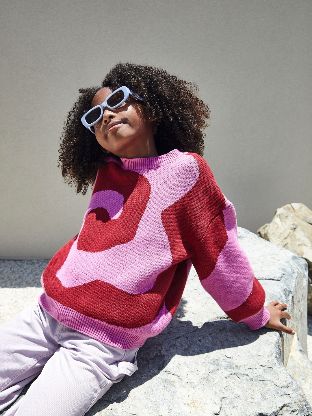 A girl wearing a kids crewneck sweater in bright pink, with a large single red swirl design that covers the entire sweater. She is seated on a rock, with a slight smile, and wears blue rectangular sunglasses.