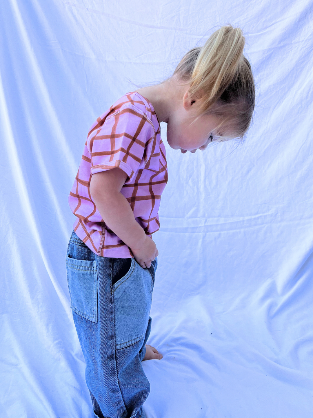 The enchanting image features a toddler in the PAINTBRUSH CHECK LAP-SHOULDER TEE, a pink and red checkered shirt, paired with oversized jeans against a crisp white backdrop. This charming scene beautifully conveys innocence and style, ideal for captivating product descriptions.