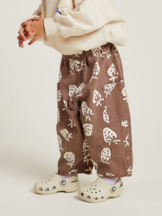 Second image of The EMERSON CHOC STRAB TROUSER are brown children's pants featuring an elastic waistband, two pockets, and a white strawberry print pattern.
