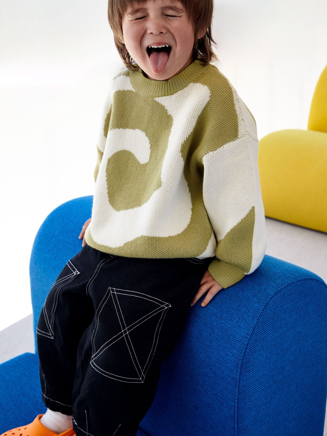 A child wearing a kids crewneck sweater in sage green, with a large single cream swirl design that covers the entire sweater. He is sticking his tongue out, with eyes closed, and sits on a blue chair. He wears black jeans with white stitching and orange shoes.