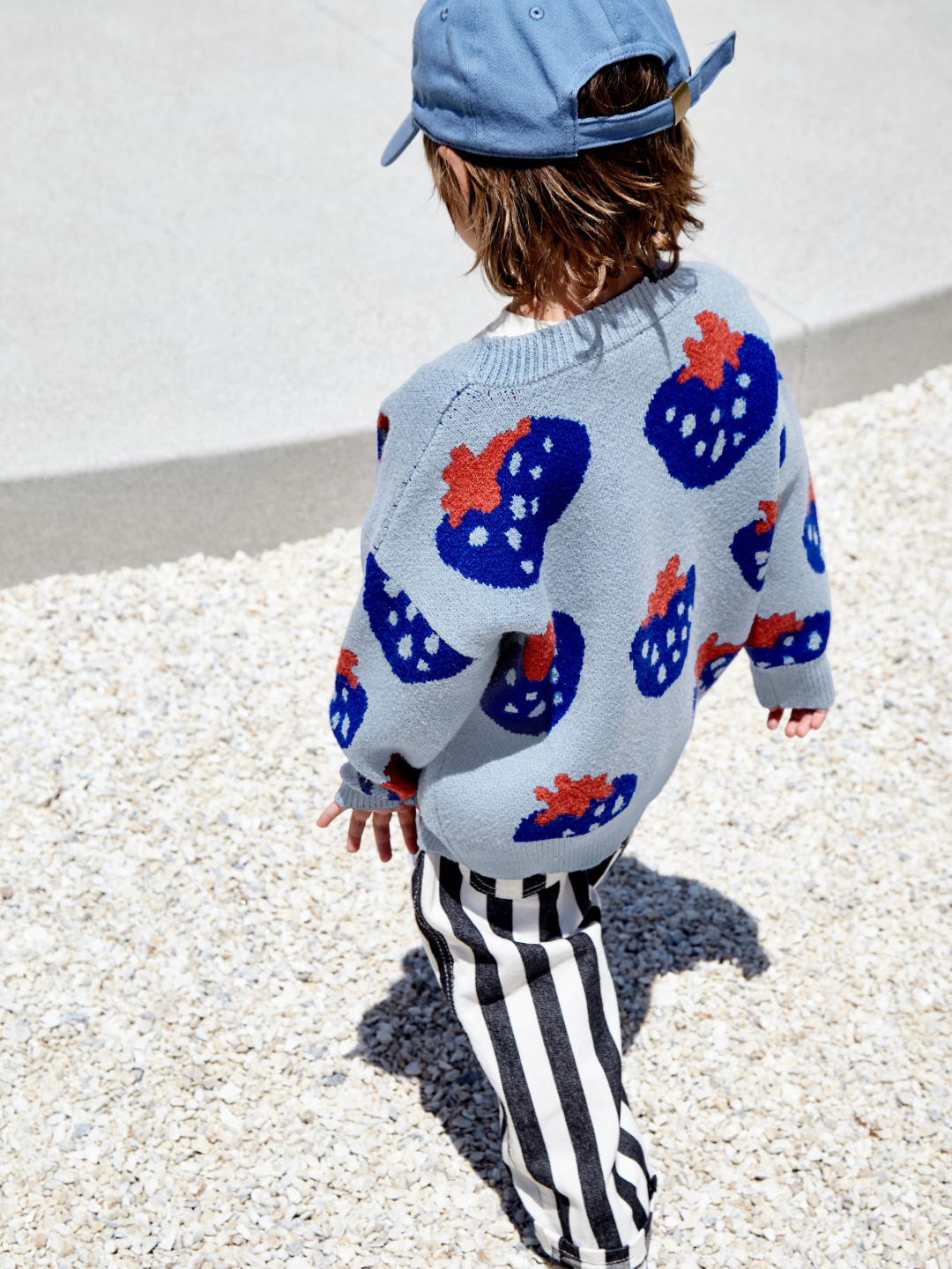 Sky | Back view of a child wearing a kids v-neck cardigan in light blue with an all-over pattern of large blue strawberries with a red leaf. He is walking on gravel in an outdoor space.