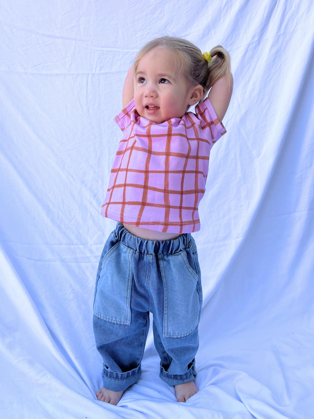 A young child with pigtails stands barefoot on a white cloth, delightfully raising her arms while wearing the PAINTBRUSH CHECK LAP-SHOULDER TEE in pink and blue jeans, creating a captivating image.