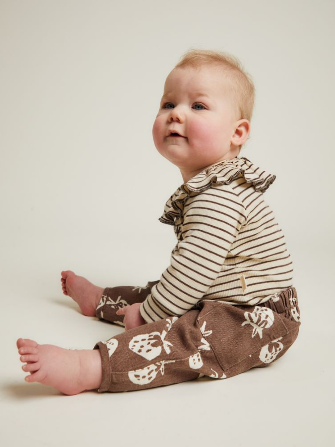 Baby sitting on the floor wearing a striped long-sleeve top with a ruffle collar and EMERSON CHOC STRAB TROUSER, looking slightly upward.
