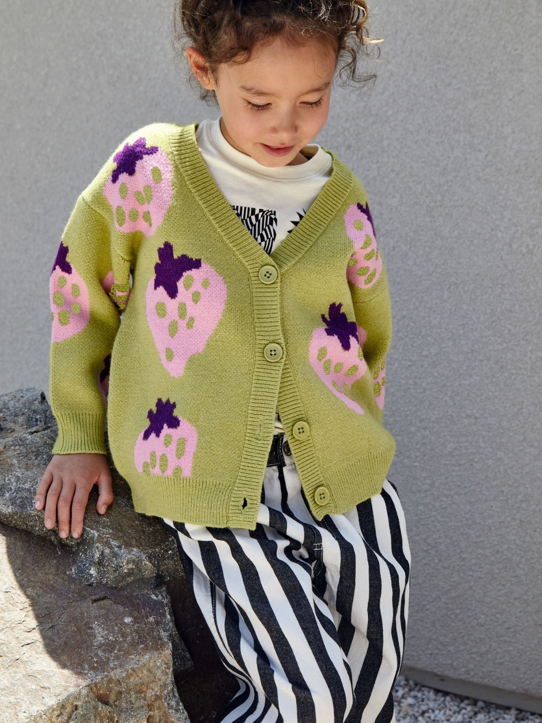 Pistachio | A child wearing a kids v-neck cardigan in pistachio green with an all-over pattern of large pink strawberries with a purple leaf. Cardigan has 4 buttons.She wears it with a white graphic tee, black and white striped jeans, and is seated on a rock against a grey wall.