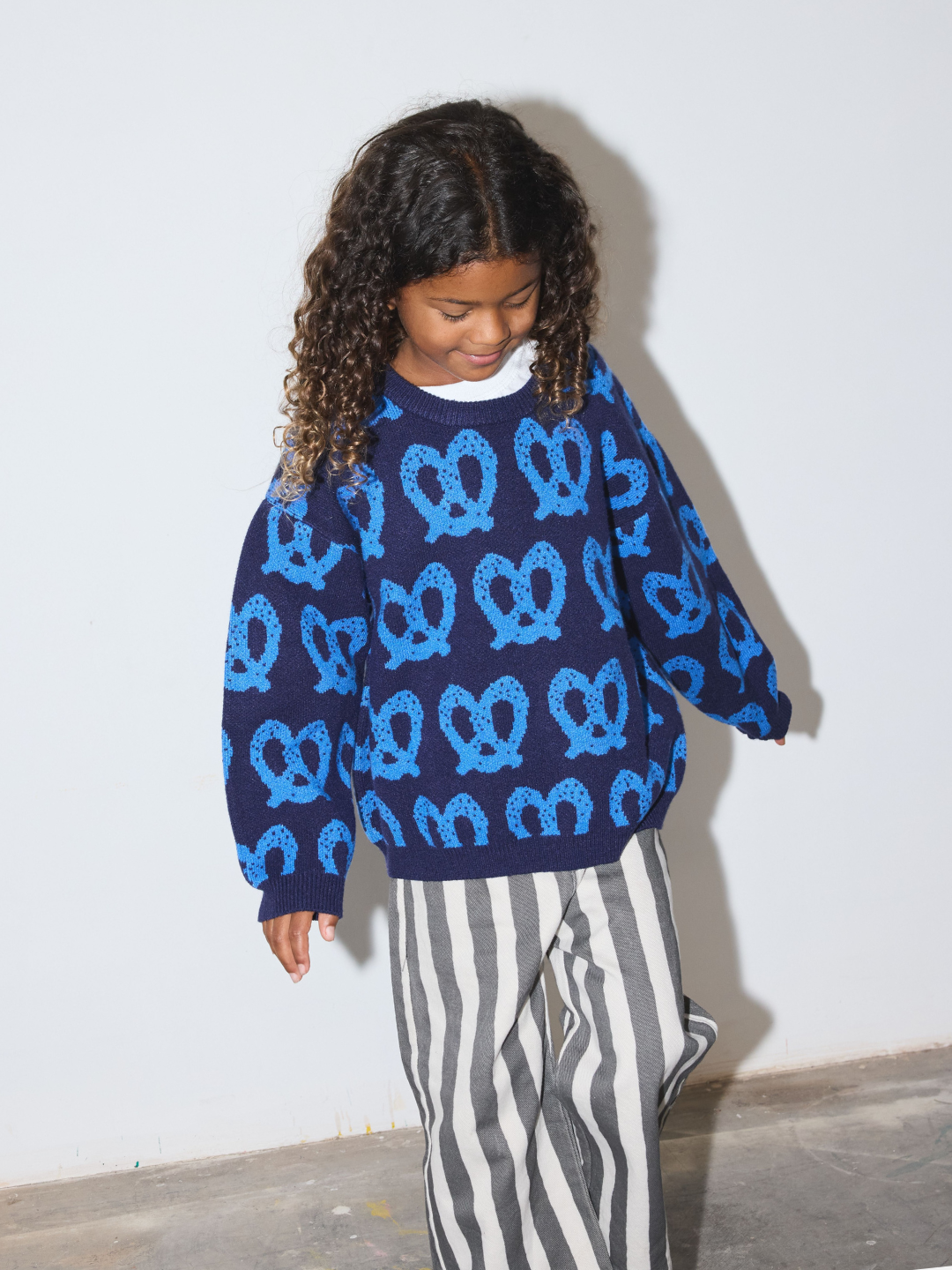 Blue | A child wearing a navy blue sweater with a repeating light blue pretzel pattern, paired with gray and white vertically striped pants, standing against a plain white wall.