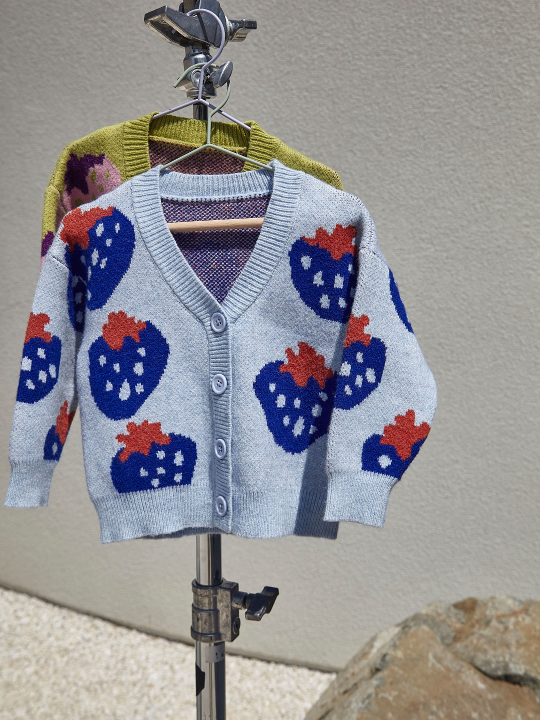 Sky | Two kids v-neck cardigans, shown on hangers, hanging from a metal photography stand, in an outdoor space with a gravel floor and grey wall. The cardigan in front is light blue with an all-over pattern of large blue strawberries with a red leaf. The cardigan behind is green with a pink strawberry with a purple leaf.
