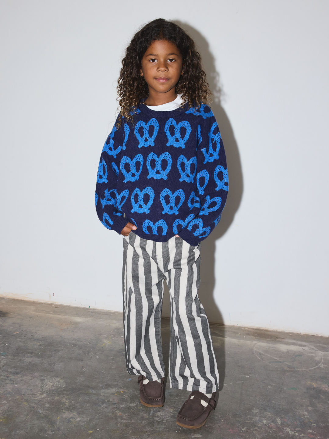 A child wearing a navy blue long-sleeve sweatshirt with a bright blue pretzel knot pattern, paired with black and white vertically striped wide-leg pants and brown shoes, standing against a plain white wall.