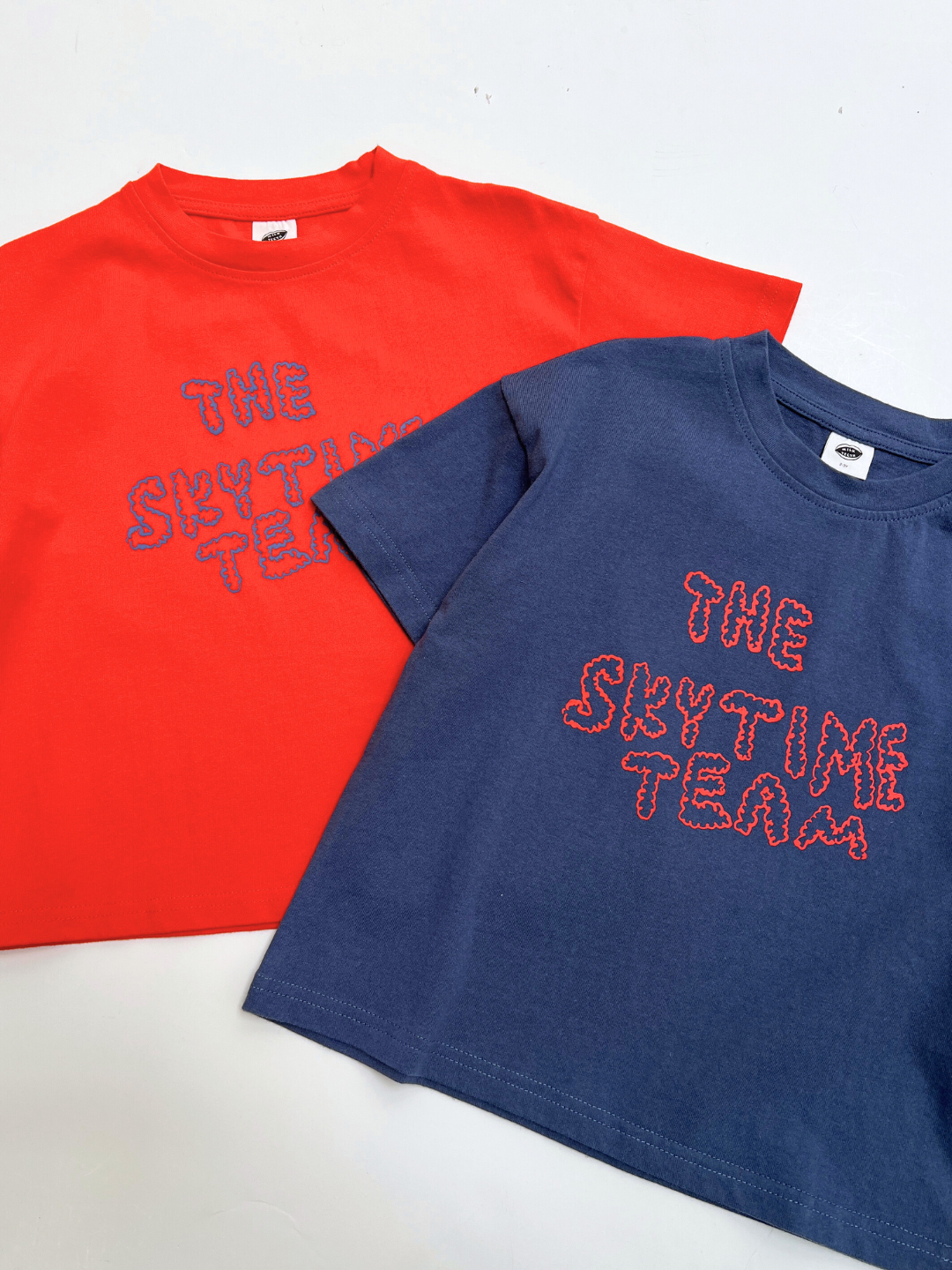 Blue | Two oversized, boxy SKYTIME TEE shirts—one red, one navy—feature "THE SKYTIME TEAM" stitched in playful lettering on the front. Both tees are artist-designed and laid flat on a white surface.