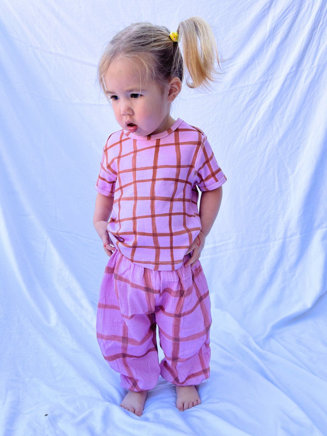 Pink/Terracotta | A young child with a ponytail, barefoot against a white backdrop, wears PAINTBRUSH CHECK MUSLIN PANTS in pink with a hand-drawn grid check pattern, displaying a thoughtful expression.