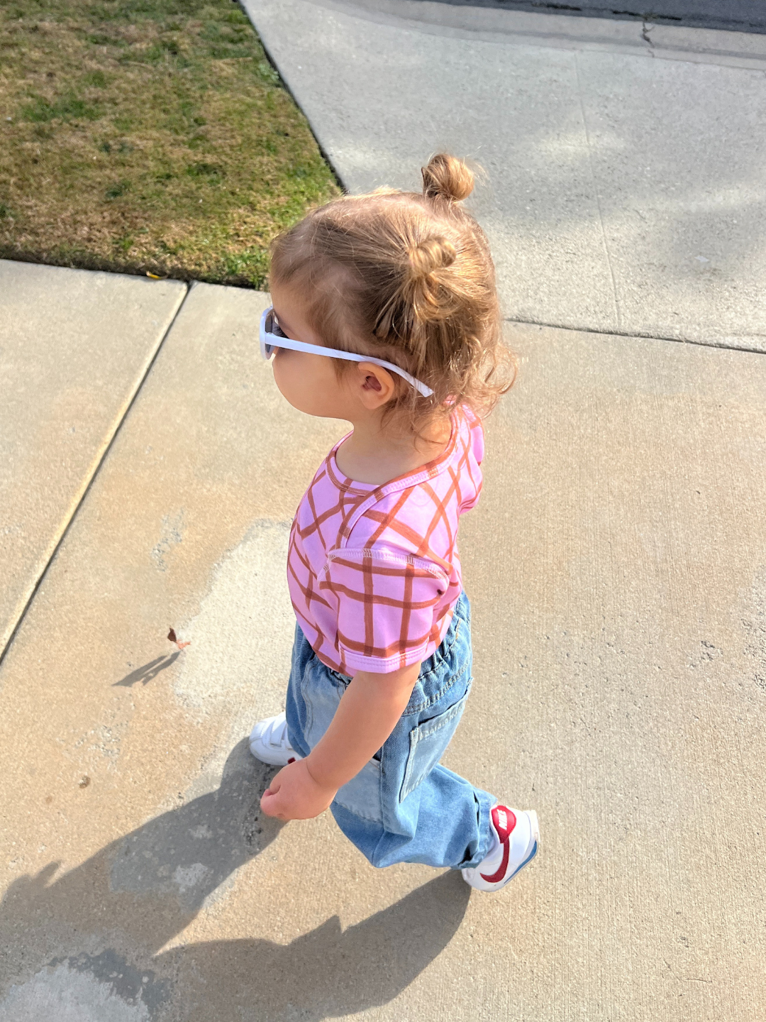 A cute toddler struts down the sidewalk in a PAINTBRUSH CHECK LAP-SHOULDER TEE, jeans, and white sneakers. With hair in a tiny bun and white sunglasses perched on their nose, this scene captures charming imagery of youthful style.